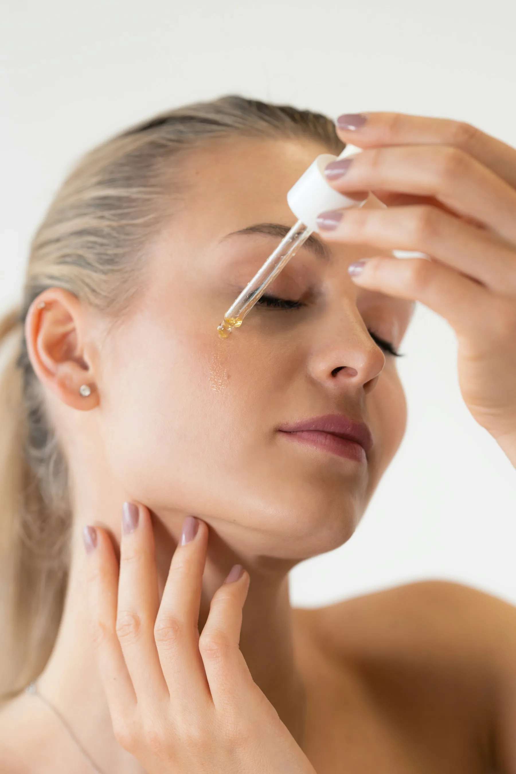 A woman with her eyes closed applying a drop of facial serum from a glass dropper